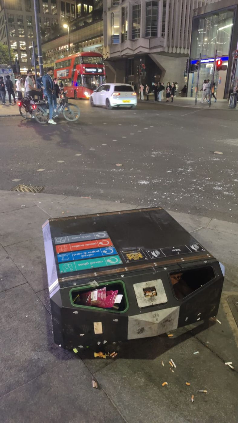 A recycling bin lying on its side across the pavement on Tottenham Court Road at night, cigarette ends and broken glass scattered around it; a red double-decker bus, the Tube station and passers-by in the background.